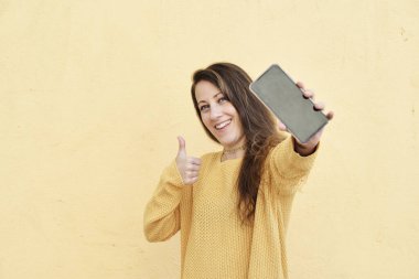 Gülümseyen mutlu bir kadın cep telefonu kullanıyor. Ekran ekranı boş. Sarı arka planda izole edilmiş bir jest gibi.