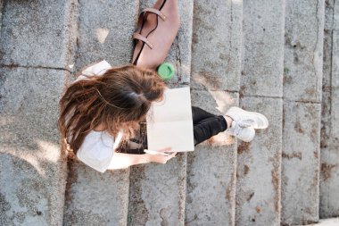 Merdivenlerde oturan bir kız öğrencinin üst görüntüsü ve bir deftere yazı yazması.