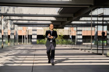 Resmi takım elbiseli Latin işadamı güneş ışığı altında modern kentsel iş bölgesinde tabletle güvenle dışarı çıkıyor.