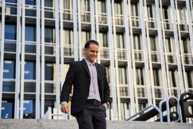 Resmi giyinmiş gülümseyen Latin iş adamı modern cam ofis binasının önünde yürüyor, başarıyı ve güveni temsil ediyor.