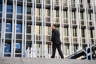 Resmi takım elbiseli Latin işadamı modern cam ofis binasının dışına çıkıyor, başarıyı ve hırsı simgeliyor.