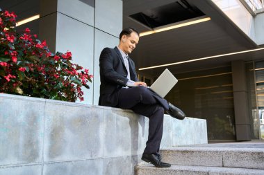 Resmi takım elbiseli Latin işadamı modern ofis binasının dışında oturuyor, kulaklıklı dizüstü bilgisayar kullanıyor ve güvenle gülümsüyor.