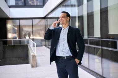 Resmi giysiler içinde gülümseyen Latin iş adamı modern ofis binasının önünde cep telefonuyla konuşuyor.