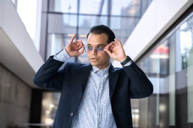 Takım elbiseli şık Latin adam güneş gözlüğünü ayarlıyor. Modern bir binanın içinde cam duvarları var.