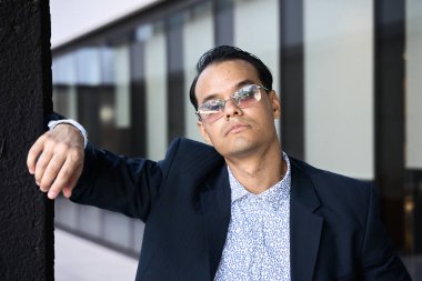 Güneş gözlüklü ve takım elbiseli şık Latin bir adam modern şehir iş ortamında duvara yaslanıyor.