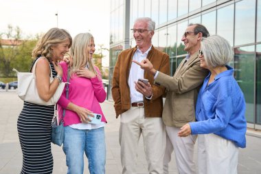 Bir grup mutlu kıdemli arkadaş cep telefonuyla bir şeyler paylaşmaktan keyif alıyor, birlikte gülüyor ve dışarıda gülüyorlar.
