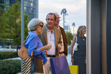 Bir grup kıdemli arkadaş alışveriş torbaları taşıyor ve şehirde gezintiden zevk alırken bir mağazanın camına bakıyor.
