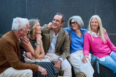 Bir grup mutlu kıdemli arkadaş gülümsüyor ve kucaklaşıyor, şehir gezisi sırasında keyifli bir anı paylaşıyorlar. Şehirde sosyalleşme ve kaynaşma