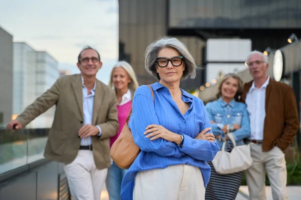Arkadaşları arka planda gülümserken kollarını kavuşturmuş, ciddi bir ifadeyle kıdemli bir kadın.