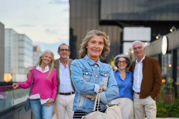 Kollarını kavuşturup kameraya gülümseyen kendinden emin yaşlı bir kadın. Arka planda bulanık bir grup olgun arkadaşla.