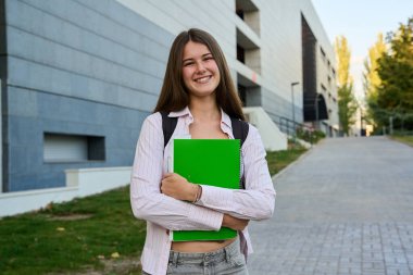 Sırt çantalı, elinde yeşil defterler olan gülümseyen genç kız bir üniversite kampüsü yolunda duruyor.