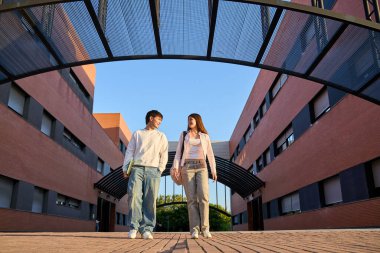 Modern üniversite binaları arasında yürüyen iki genç öğrenci, açık bir gökyüzünün altında kitap ve sırt çantaları taşıyorlar.