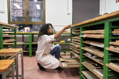 Stüdyo atölyesindeki rafları inceleyen ve düzenleyen kadın sanatçı, özel bir parça arıyor.