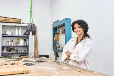 Laboratuvar önlüğü giymiş gülümseyen bir kadın temiz bir atölyede duruyor, kollarını kavuşturuyor ve kameraya bakıyor.