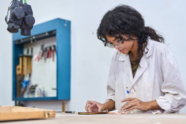 Laboratuvar önlüklü ve gözlüklü bir kadın. Aletlerle çalışıyor. Parlak bir atölyedeki tezgahta metal mücevher parçaları yapıyor.