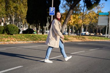 Güneşli bir sonbahar gününde şehrin caddesinden mutlu bir şekilde geçen genç bir kadın, şehir ortamında gündelik yürüyüşünün keyfini çıkarıyor.
