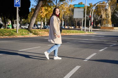 Genç bir kadın güneşli bir sonbahar gününde güvenli bir şekilde caddeyi geçiyor, gülümsüyor ve şehir hayatının tadını çıkarıyor.