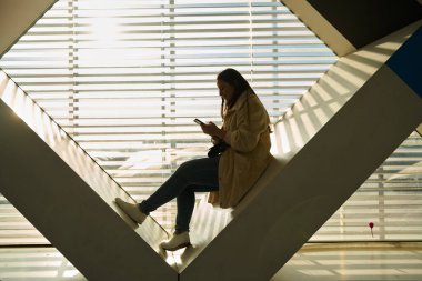 Pencerenin kenarında oturan kadın, akıllı telefondan mesaj atıyor, güneş ışığı duvarda geometrik gölgeler yaratıyor.