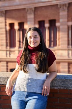 Esmer kadın mutlu bir ifadeyle poz veriyor, taş bir duvara yaslanıyor ve arka planda eski bir tuğla bina var.