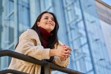 Genç bir kadın gülümsüyor, modern bir şehir binasına bakıyor, umut, mutluluk ve gelecek için olumlu bir görünüm ifade ediyor.