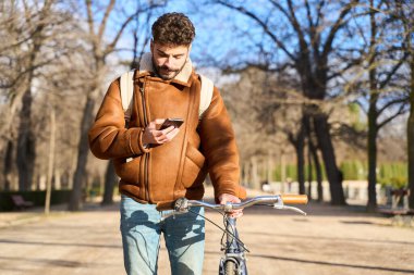 Genç adam dışarıda duruyor, akıllı telefonunu kontrol ediyor ve bisiklet gidonu tutuyor, şehir yaşam tarzını ve bağlantısını temsil ediyor.