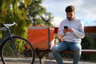 Bisikletli kurye olarak çalışan genç adam parkta dinlenirken yeni siparişler için akıllı telefonunu kontrol ediyor.