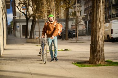 Yeşil kasklı ve sırt çantalı bir adam bisikletini güneşli bir şehir kaldırımında ağaçların ve park edilmiş arabaların önünden itiyor.