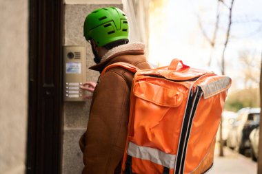 Kasklı ve sırt çantalı bir teslimatçı dahili telefon düğmesini çalıyor ve şehir sakinlerine önemli hizmetler sunuyor.