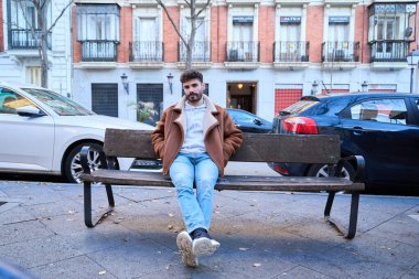 Şehir sokağındaki ahşap bir bankta rahat bir şekilde oturan genç adam, sıradan bir kış ceketi ve kapüşonlu bir şey giyiyor.