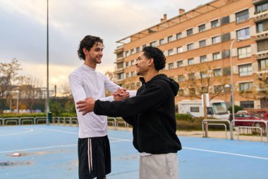 Bir basketbol sahasında birbirine bağlanmış gülümseyen iki adam. Birbirlerini tokalaşarak selamlıyorlar. Arkadaşlık ve aktif yaşam tarzlarını gösteriyorlar.
