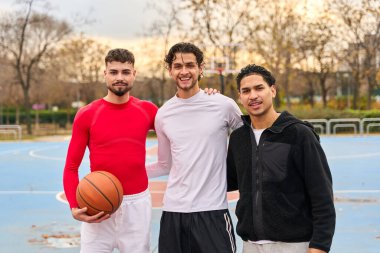 Açık hava basketbol sahasında duran üç gülümseyen genç adam, arkadaşlığı, şehir sporunu ve gençlik kültürünü temsil ediyor.