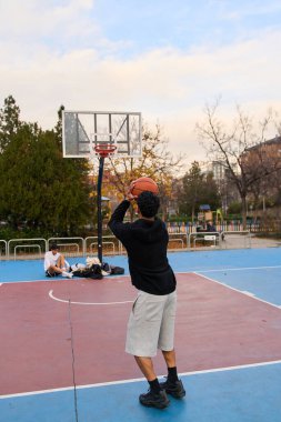 Şehir parkında basketbol oynayan genç bir adam. Bir başkası ise sonbahar ağaçları ve binalarla çevrili kenarda dinleniyor.