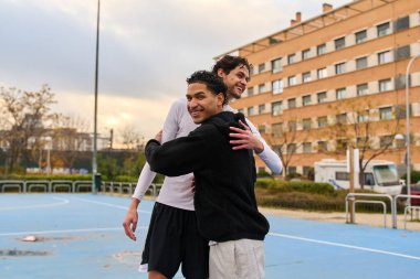 Mavi bir basketbol sahasında gülümseyen ve sarılan iki genç erkek arkadaş spor ve bağlantıyı kutluyorlar.