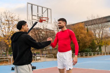 Açık hava basketbol sahasında el sıkışan iki genç adam takım çalışması, dostluk ve sportmenliği temsil ediyor.