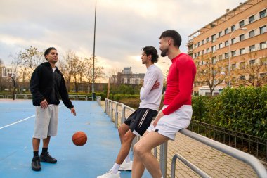 Şehirdeki bir basketbol sahasında antrenman yapan ve dinlenen üç genç adam sıradan bir spor aktivitesinin keyfini çıkarıyorlar.