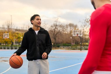 Genç adam mavi sahada turuncu top sürüyor, açık hava antrenmanının keyfini çıkarırken arkadaşına bakıyor.