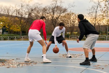 Açık bir sahada basketbol oynayan üç adam top sürüyor ve sporla meşgul oluyor.