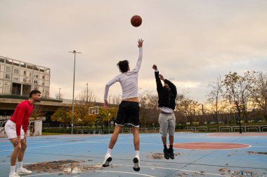 Atletik gençler aktif bir oyun sırasında canlı bir mavi şehir sahasında atlayıp basketbol için yarışıyorlar.