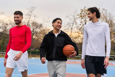 Çeşitli arkadaşlar gülümsüyor, konuşuyor ve dışarıda bir sahada basketbol topu taşıyor, spor ve arkadaşlıktan keyif alıyorlar.