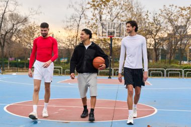Üç genç adam basketbol sahasında yürüyor, biri top taşıyor. Arkadaşlar dışarıda kaynaşıyor, oyun için hazır.