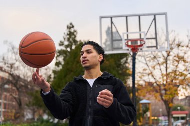 Kendine güvenen genç bir adam parmağında basketbol topunu dengeliyor, şehir basketbol sahasında bir numara yapıyor.