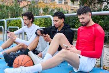 Çeşitli erkek arkadaşlar cep telefonlarıyla çevrimiçi olarak bir basketbol sahasında oturuyorlar.