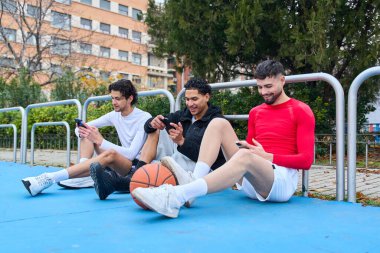Üç arkadaş basketbol sahasında rahatlıyor, akıllı telefonlar kullanıyor, gençlik yaşam tarzını, şehir sporunu ve bağlantıyı temsil ediyor.