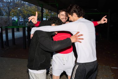 Üç genç erkek sporcu, spor yaptıktan sonra sıkı bir dostluk ve takım çalışması göstererek dışarıda sarılıp kutluyor.