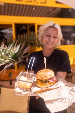 Restoran personeli, güneşli bir terasta patates dilimli gurme domuz burger servis ederek menüler, markalaşma ve gıda pazarlaması için ideal bir dostça bir misafirperverlik atmosferi yaratıyor..