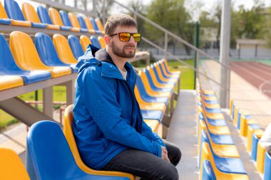 Stadyum koltuğunda oturan güneş gözlüklü ve mavi ceketli bir adam. Stadyum koltukları sarı ve mavi..