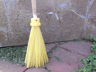 Tahta saplı parlak sarı bir plastik süpürge taş duvarın karşısında duruyor, yanında biçilmiş çimenler var..