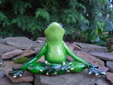 Komik şirin bir kurbağa (biblo, oyuncak) bahçede kaydırak üzerinde oturuyor ve bitkilerin arka planına karşı yoga yapıyor..