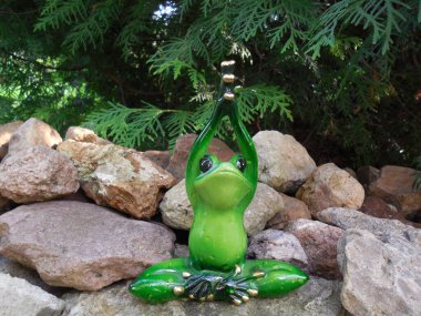 Komik şirin bir kurbağa (biblo, oyuncak) bahçede kaydırak üzerinde oturuyor ve bitkilerin arka planına karşı yoga yapıyor..