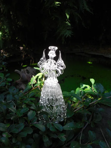 A forest fairy or an angel (a decorative figure made of glass, a souvenir, the property of the author of the photo) by the pond. In the shade, bright sunlight penetrates through the branches.